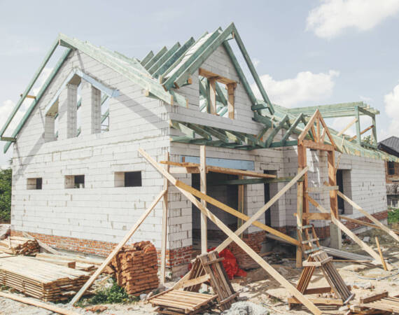 Unfinished modern farmhouse building. Wooden roof framing of mansard with dormer and aerated concrete block walls with windows. Rafters and beams on blocks. New house construction